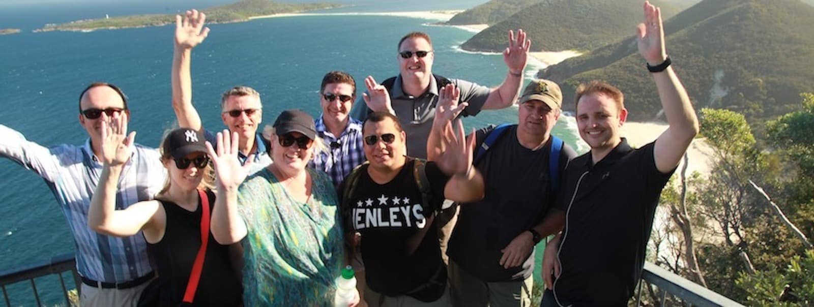 Nine people raise their hands and make smiles with a spectacular sight of landscape behind them.
